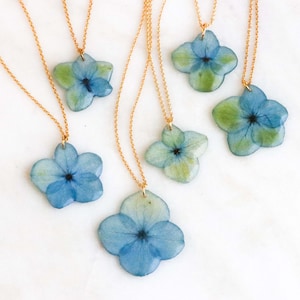 Peut inclure: Colliers à chaîne dorée avec des pendentifs représentant des fleurs d'hortensia bleues et vertes pressées. Les pendentifs floraux ont un revêtement en résine transparente. Les colliers sont présentés sur une surface en marbre blanc.
