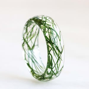 May include: A clear resin ring with green grass embedded inside. The ring is thick and has a rounded shape.