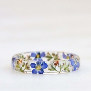May include: A clear resin ring with embedded blue and white flowers. The ring is a delicate and unique piece of jewelry.