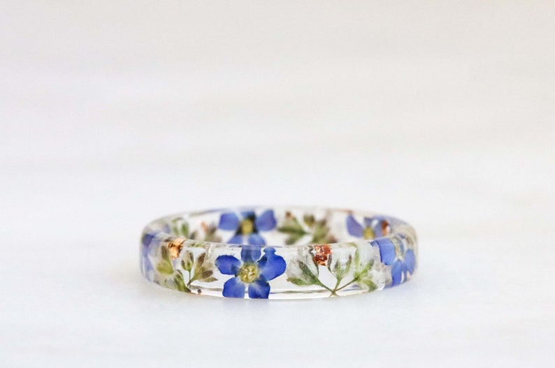 May include: A clear resin ring with embedded blue and white flowers. The ring is a delicate and unique piece of jewelry.