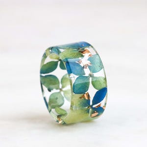May include: A clear resin ring with embedded blue and green pressed flowers and gold leaf accents.