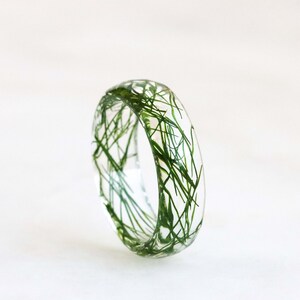 May include: A clear resin ring with green grass embedded inside. The ring is a simple band with a smooth, rounded surface.