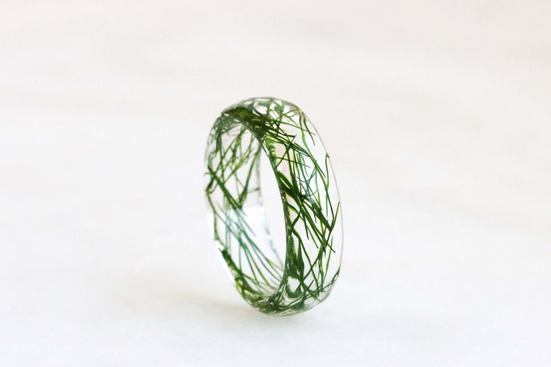 May include: A clear resin ring with green grass embedded inside. The ring is a simple band with a smooth, rounded surface.