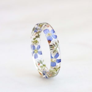 May include: A clear resin ring with embedded blue and white flowers. The ring is a delicate and unique piece of jewelry.