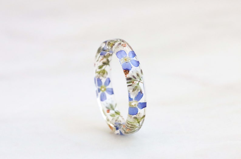 May include: A clear resin ring with embedded blue and white flowers. The ring is a delicate and unique piece of jewelry.