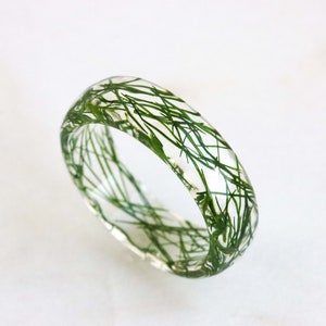 May include: A clear resin ring with green dried plant material embedded inside. The ring is thick and has a rounded shape.