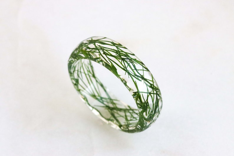 May include: A clear resin ring with green dried plant material embedded inside. The ring is thick and has a rounded shape.
