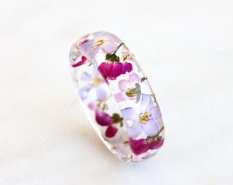 May include: A clear resin bangle bracelet featuring dried flowers in shades of purple, pink, and white. The flowers are set within the transparent bracelet, creating a unique floral accessory. The bracelet is a stylish piece of jewellery.
