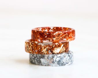 Könnte beinhalten: Drei Harzringe übereinander gestapelt. Der oberste Ring ist orange mit Kupferflocken. Der mittlere Ring ist bernsteinfarben mit Goldflocken. Der untere Ring ist grau mit Silberflocken. Jeder Ring ist aus transparentem Harz gefertigt.