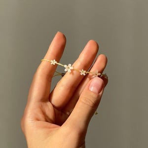 Könnte beinhalten: Ein zartes Armband mit kleinen weißen Blumenanhängern, die auf einer dünnen, hellgelben Kordel aufgereiht sind. Das Armband wird in einer Hand vor einem neutralen grauen Hintergrund gehalten und zeigt sein schlichtes, elegantes Design. Dieses Accessoire ist perfekt.