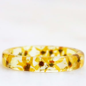 May include: A clear resin ring with yellow flowers pressed inside. The ring is a light yellow color and the flowers are a darker yellow.