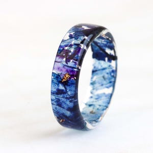 Op de afbeelding: Een harsring met een gemarmerd ontwerp in blauwe en paarse tinten, met gouden vlekken. De ring is een bandstijl, met een heldere, glanzende afwerking. De ring staat tegen een witte achtergrond.