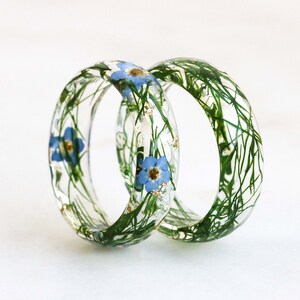 May include: Two clear resin rings with embedded blue flowers and green foliage. The rings are stacked on top of each other.