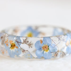 Transparent resin ring with pressed blue forget-me-not flowers, delicate gypsophila buds, and metallic flakes. Polished, lightweight design. Faceted, diamond shape.