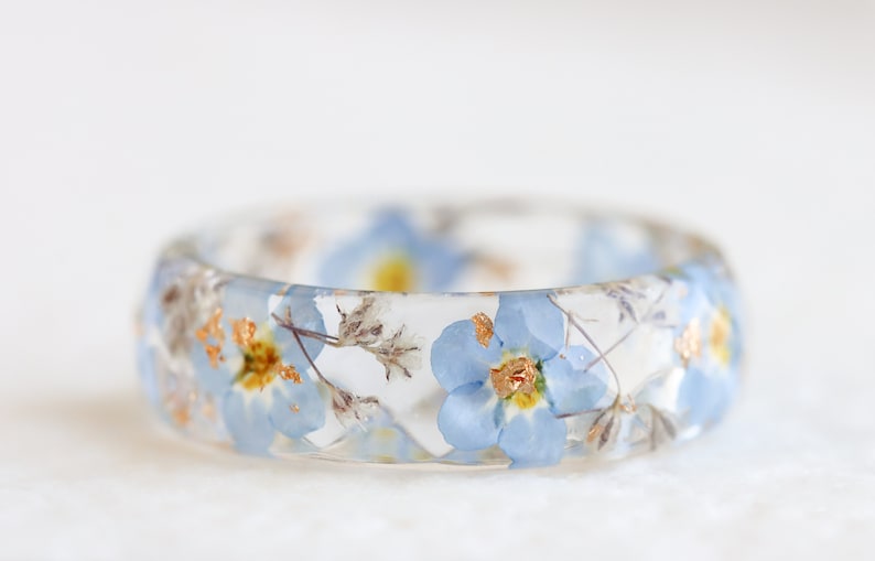 Transparent resin ring with pressed blue forget-me-not flowers, delicate gypsophila buds, and metallic flakes. Polished, lightweight design. Faceted, diamond shape.