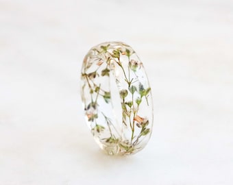 May include: A clear resin ring with dried flowers and leaves embedded inside. The ring is a light brown color and has a smooth, polished finish.