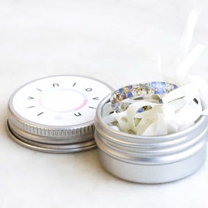 May include: A silver metal tin with a white lid. The lid has the word "INIOVE" printed on it. The tin contains a blue and white patterned ring.