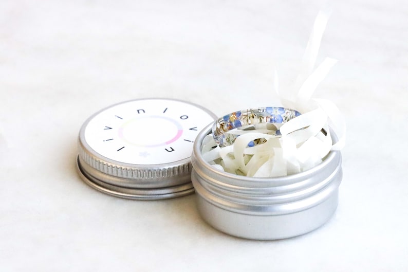 May include: A silver metal tin with a white lid. The lid has the word "INIOVE" printed on it. The tin contains a blue and white patterned ring.