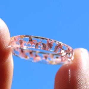 以下が含まれることがあります： 透明な樹脂製のリングで、バンド全体に小さなピンク色の花びらが埋め込まれています。円形のバンドは透明で、光が素材を透過し、樹脂の中に閉じ込められた自然なピンク色の花のディテールを際立たせています。