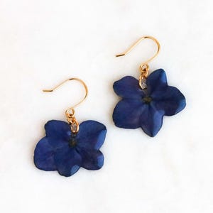 May include: A pair of gold-coloured drop earrings with dark blue, flower-shaped pendants. The earrings are set against a white background, highlighting the contrast between the gold and the deep blue of the floral design.