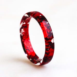 Peut inclure: Une bague en résine rouge et blanche. La bague est une bande avec une finition brillante. La couleur rouge présente des variations sombres et claires, avec des accents blancs. La bague est posée sur un fond blanc.