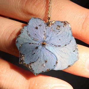 May include: A pendant necklace featuring a preserved light blue hydrangea flower petal encased in clear resin with embedded gold leaf flakes, suspended from a thin gold-toned metal chain.
