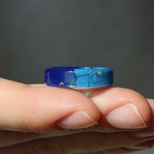 May include: A blue resin ring with gold flakes. The ring features a gradient of dark blue to light blue. The ring is held in a hand, showcasing its design and color variations. A unique piece of jewelry.