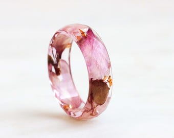 May include: A clear resin ring with embedded pink and brown dried flowers and gold flakes. The ring is a solid band, showcasing the delicate floral elements suspended within the transparent material. The ring is a unique piece of jewellery.
