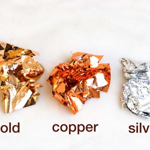 May include: Three crumpled pieces of metallic leaf, gold, copper, and silver, on a white background.
