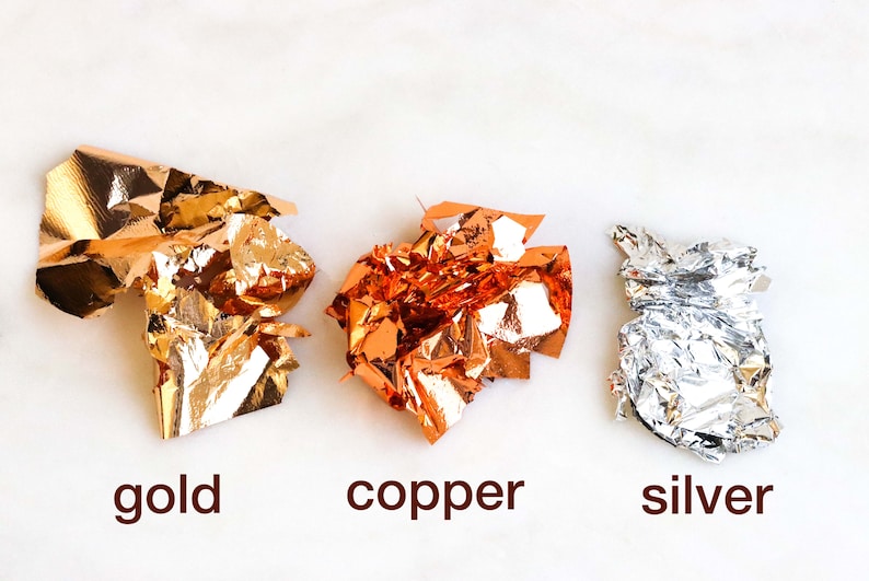 May include: Three crumpled pieces of metallic leaf, gold, copper, and silver, on a white background.