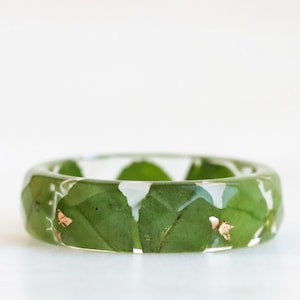 Op de afbeelding: Een ring van helder hars met groene bladeren en gouden vlokken erin.
