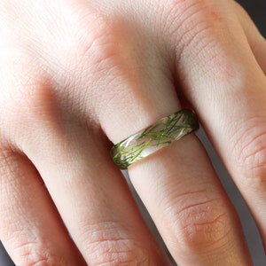 May include: A clear resin ring with green plant material embedded inside. The ring is worn on a person's finger.
