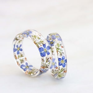 May include: Two clear resin rings with embedded blue and white flowers. The rings are stacked on top of each other.