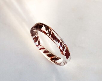 May include: A clear resin ring with embedded reddish-brown leaf-like inclusions. The ring is circular with a square profile and is set against a white background. The light catches the resin, creating highlights and shadows.
