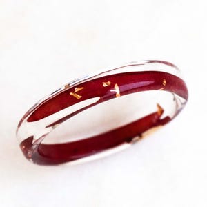 Puede incluir: Un anillo de resina transparente con una banda central de color rojo oscuro y motas doradas. El anillo está sobre un fondo blanco, resaltando su diseño transparente y colorido. Una pieza de joyería única.