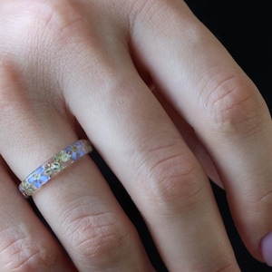 May include: A delicate ring with a band of clear resin embedded with tiny blue and green flowers.