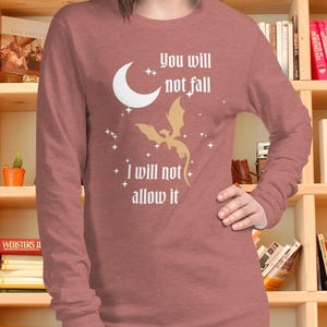 May include: A dusty rose long-sleeve shirt featuring a white graphic of a crescent moon, a flying dragon, and small stars. The text reads, "You will not fall, I will not allow it." The shirt has a crew neckline and a standard fit, made of a soft fabric.