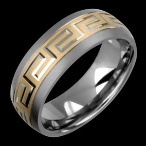 Könnte beinhalten: Silberfarbener Ring mit einem goldfarbenen griechischen Schlüsselmuster. Der Ring hat eine polierte, reflektierende Oberfläche und ein mattes Band. Das Design ist ein sich wiederholendes geometrisches Muster.