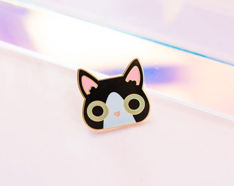 May include: Black and white enamel pin featuring a cartoon cat face with green eyes and pink ears.