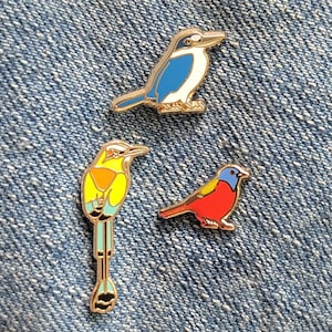 Op de afbeelding: Drie kleurrijke vogel emaille pins. Eén pin toont een blauwe en witte vogel, een andere een gele, oranje en blauwe vogel, en de derde een rode, gele en blauwe vogel. De pins hebben goudkleurige metalen randen.