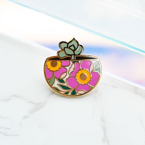 May include: A gold-colored enamel pin in the shape of a planter pot. The pot features pink flowers with yellow centers and green leaves. A small green succulent sits atop the pot. The pin is set against a white and iridescent background.