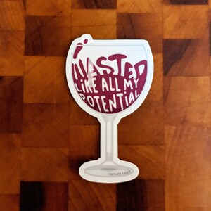 May include: White wine glass sticker with a red wine fill and the text "Wasted like all my potential" in a white outline. The sticker has a small signature at the bottom that reads "Taylor Innit".