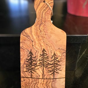 Può includere: Un piccolo tagliere rettangolare in legno di ulivo con un manico. Il tagliere presenta una venatura del legno naturale e un disegno di tre pini bruciati sulla superficie.