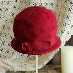 May include: A vintage cloche hat in a deep red hue, featuring a rounded crown and a small brim. The hat is adorned with a matching ribbon and a delicate veil. It is displayed on a white metal stand with floral accents, set against a rustic wooden surface.