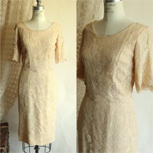 May include: A vintage, pale peach-colored lace dress with short sleeves and a knee-length skirt. The dress is displayed on a mannequin, showcasing its intricate floral lace pattern. The dress has a scoop neckline and a fitted bodice.