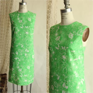 May include: A sleeveless, knee-length dress in a vibrant green hue, adorned with a white floral pattern. The dress is displayed on a vintage dress form, showcasing its classic silhouette and design. The fabric appears to be a lightweight material.