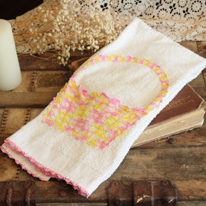 May include: White hand towel with a basket design in pink and yellow. The towel has a pink crocheted edge. A white candle and a vintage book are also in the image.