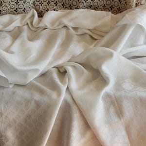 May include: A white fabric with a subtle floral design, draped and folded. The fabric appears soft and has visible shadows. A lace textile and wooden surface are visible in the background. The overall look is delicate and elegant.