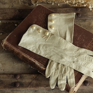 May include: A pair of vintage cream-colored leather gloves with pearl button details, resting on a worn, brown leather-bound book. The gloves are elegantly styled, suggesting a classic or antique aesthetic.