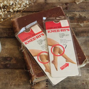 May include: Two packets of vintage Knee-Hi's stockings with red and white labels, showing illustrations of legs. The packets rest on a worn, brown book. A white candle and dried flowers are also in the image.